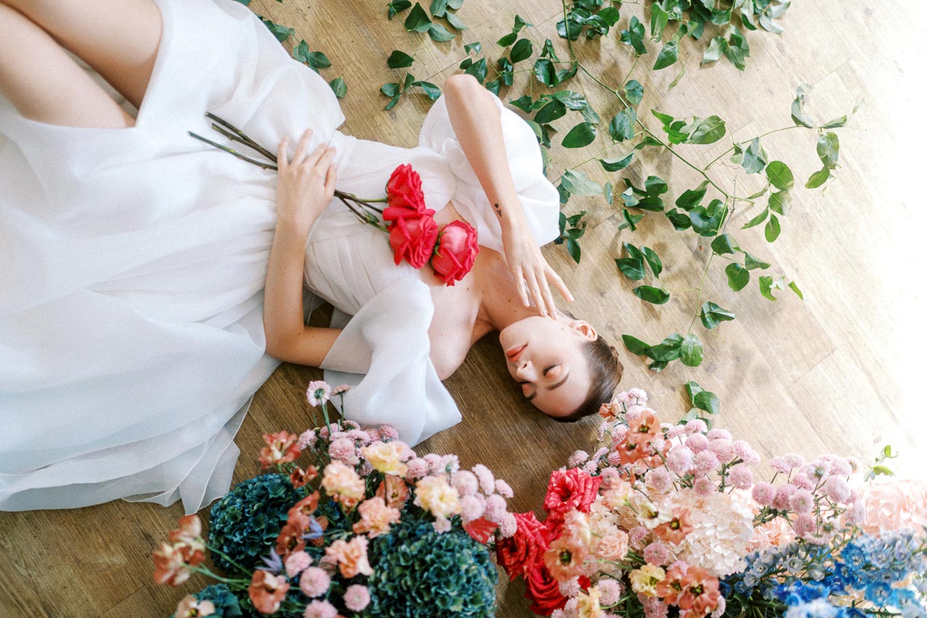A woman in a white dress lying on a wooden floor surrounded by green leaves and colorful flowers, holding pink roses.