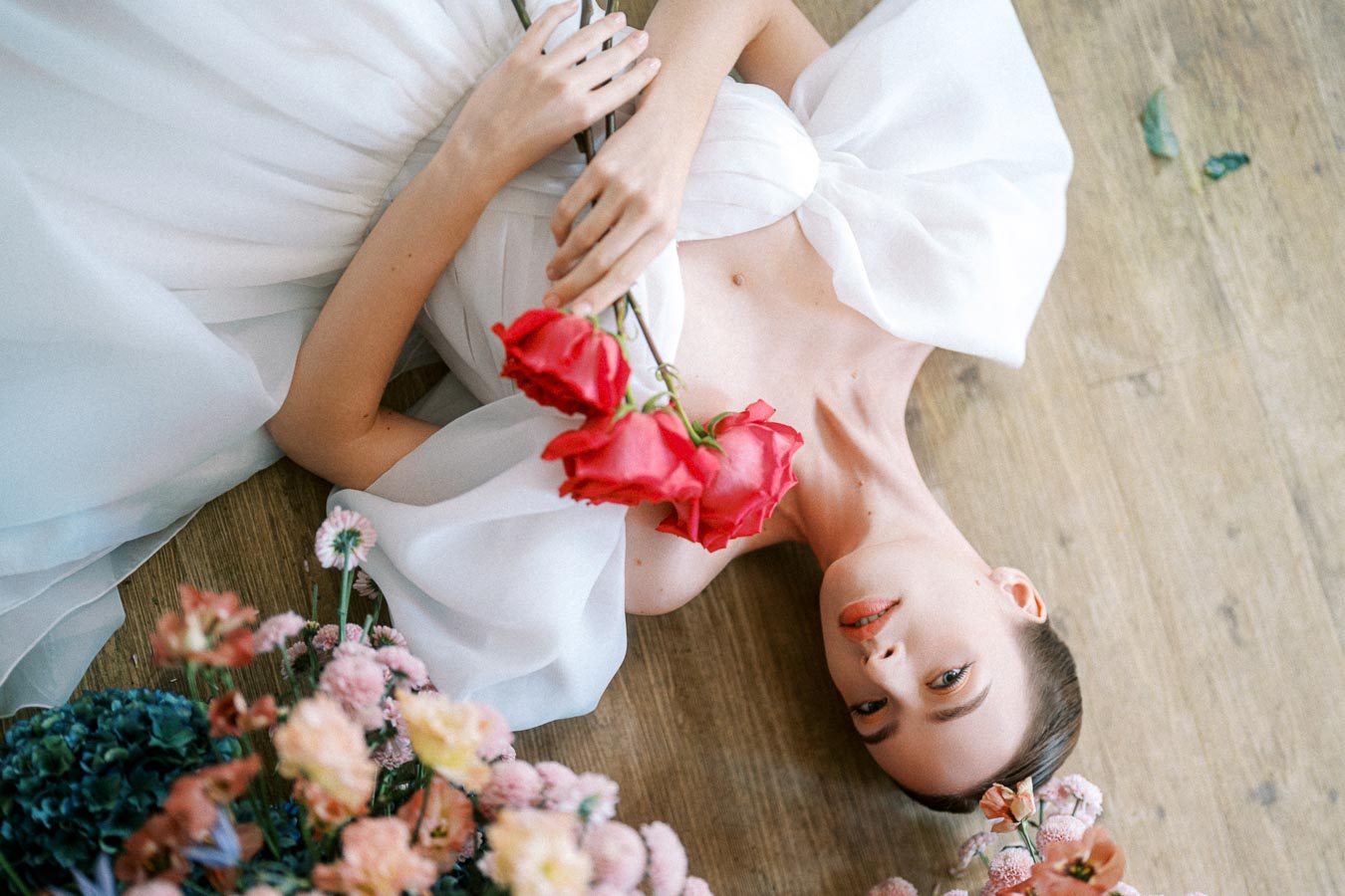 Bride in an elegant white gown holding red roses, surrounded by colorful flowers on a wooden floor background.