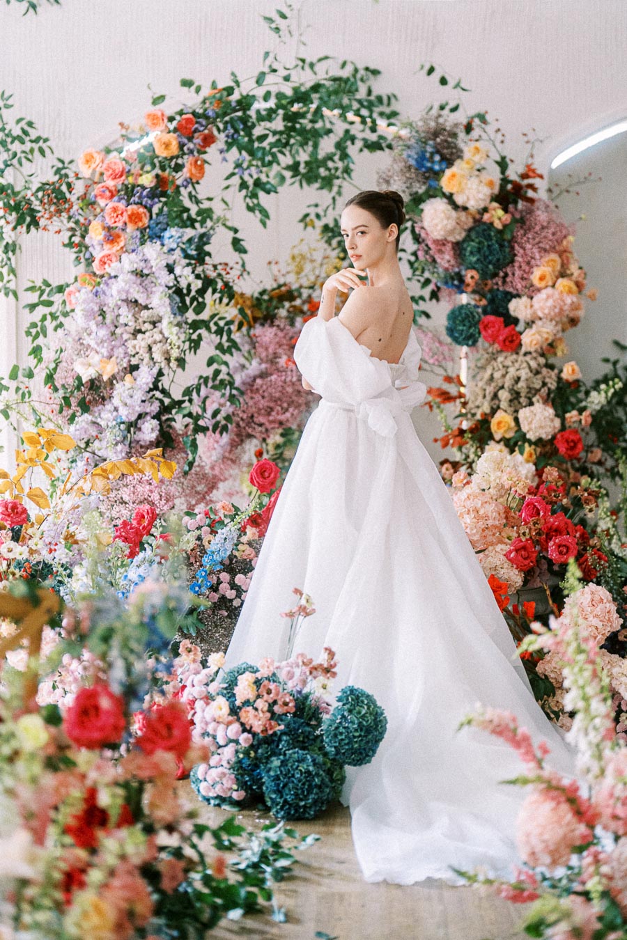 Bride in a white gown surrounded by vibrant floral arrangements, featuring roses and greenery, creating a romantic and elegant wedding scene.