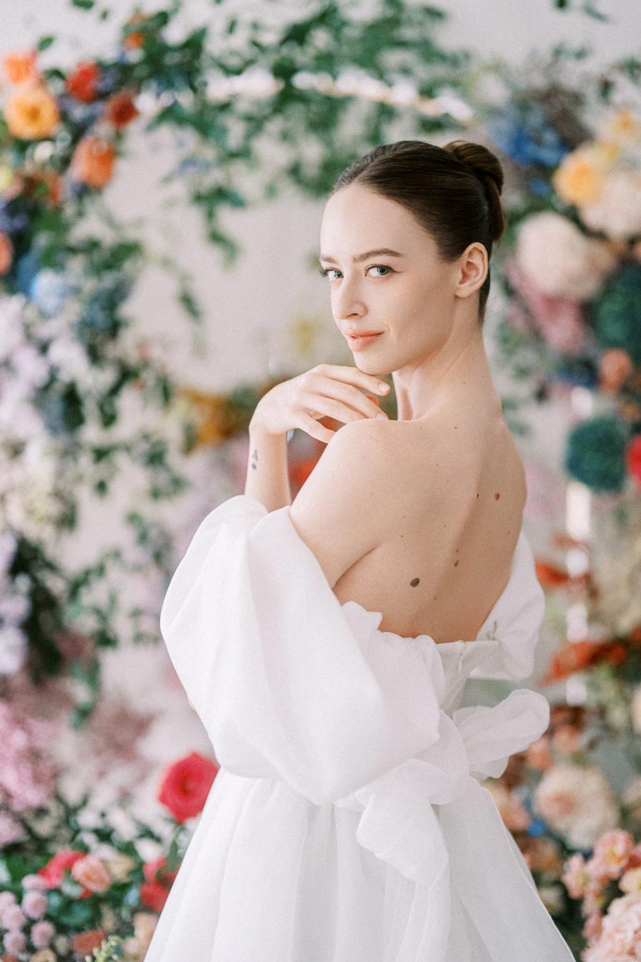 Young woman in a white off-shoulder gown poses gracefully amid a vibrant floral backdrop, capturing elegance and style.