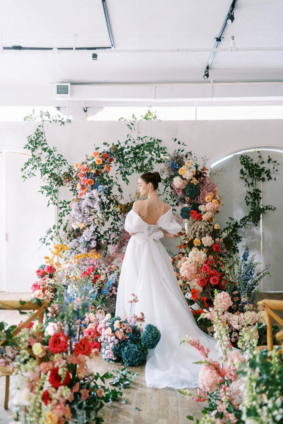 Elegant bride in a white gown standing amidst colorful floral arrangements in a bright, modern venue; showcasing wedding fashion and floral decor.
