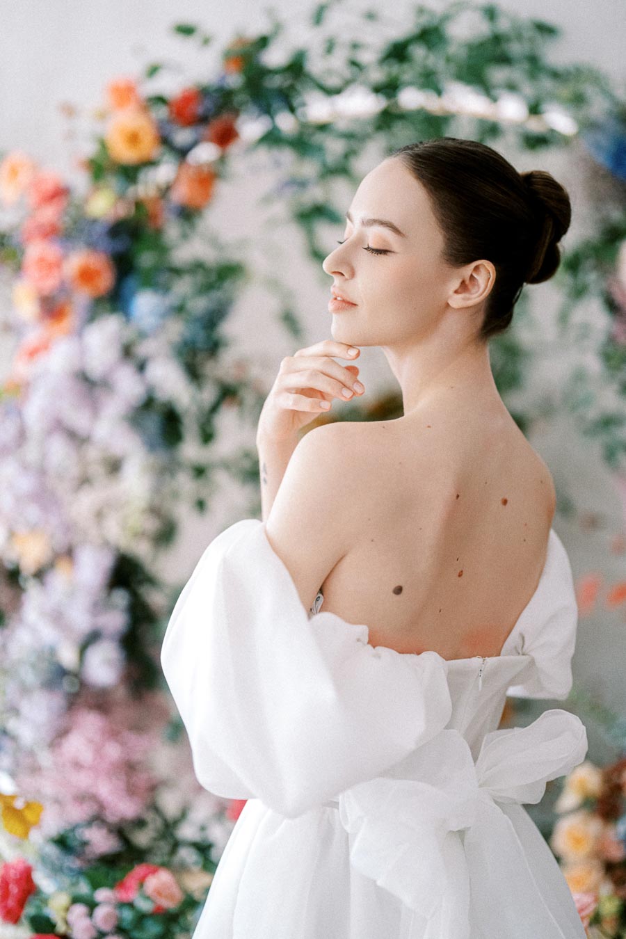 Young woman in a white, off-shoulder dress, standing against a vibrant floral backdrop, looking serene and elegant with her hair in a neat bun.