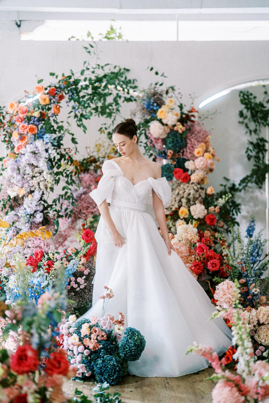 A bride in an elegant white gown surrounded by vibrant floral arrangements, featuring colorful roses and greenery, creating a romantic wedding setting.