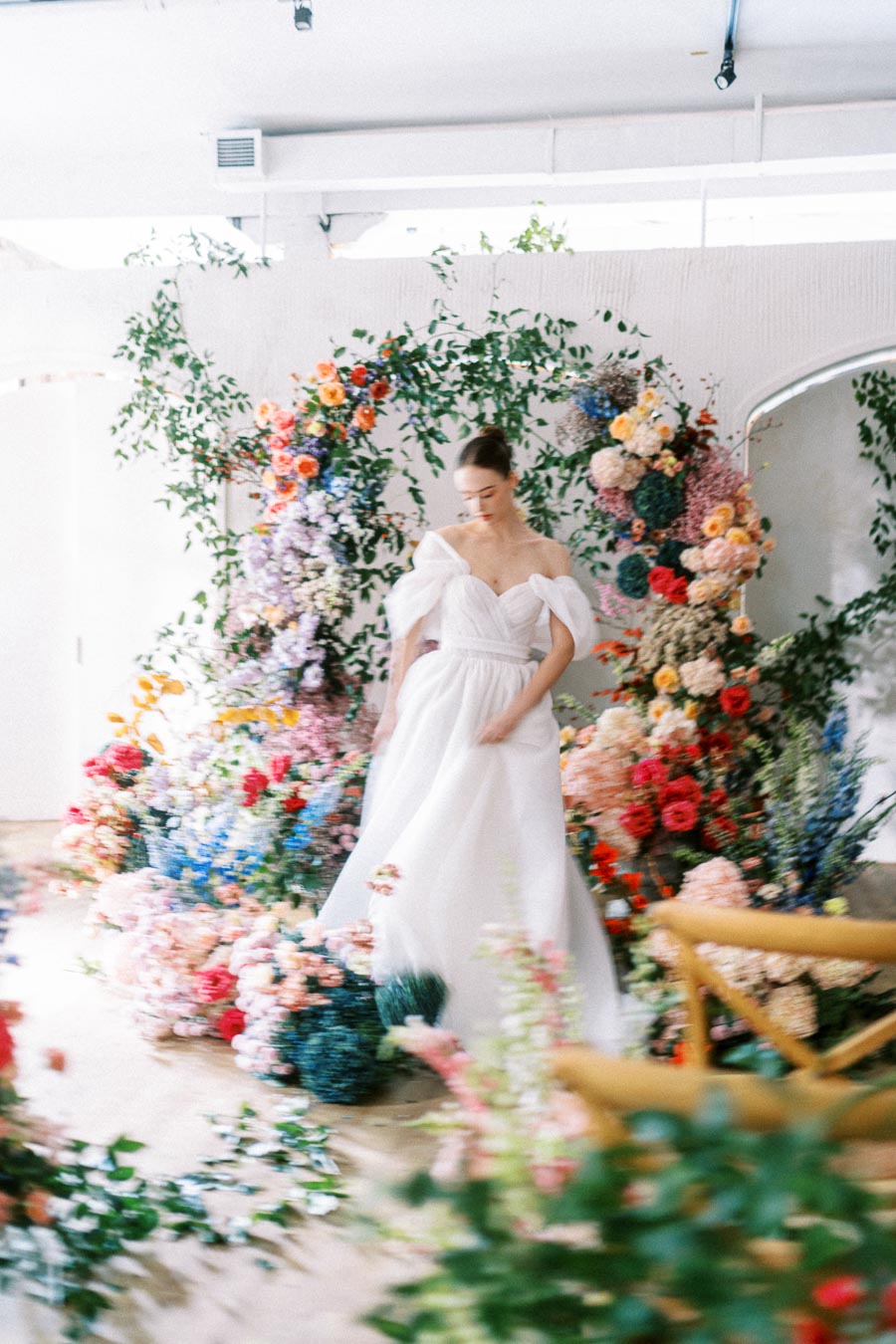 A bride in a flowing white gown stands gracefully in a room filled with vibrant, colorful floral arrangements, creating a romantic and whimsical wedding scene.