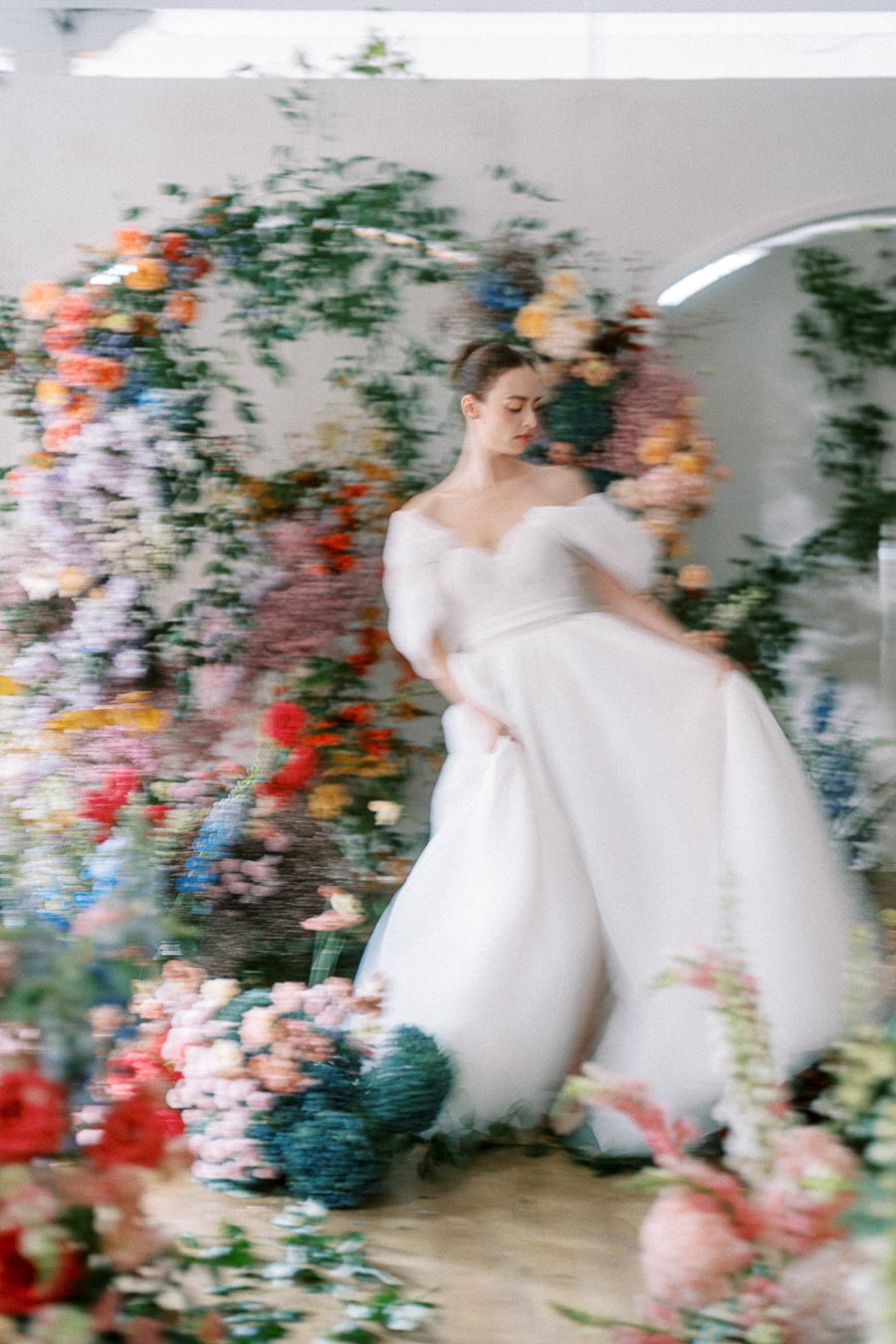 Blurry artistic photo of a bride in an elegant white wedding dress surrounded by vibrant floral arrangements.