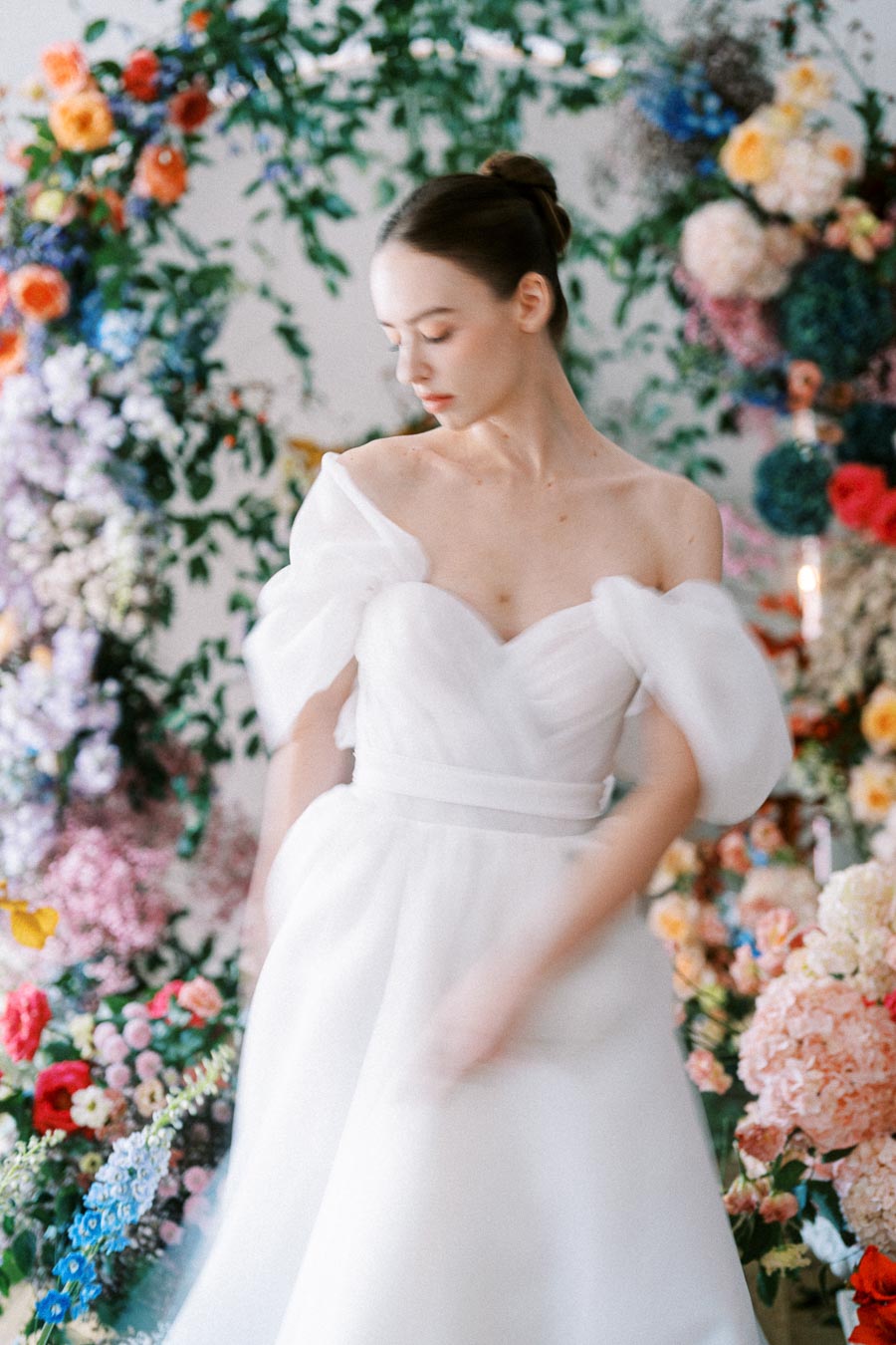 Elegant bride in a flowing white wedding dress against a backdrop of vibrant, multicolored floral arrangements and lush greenery.