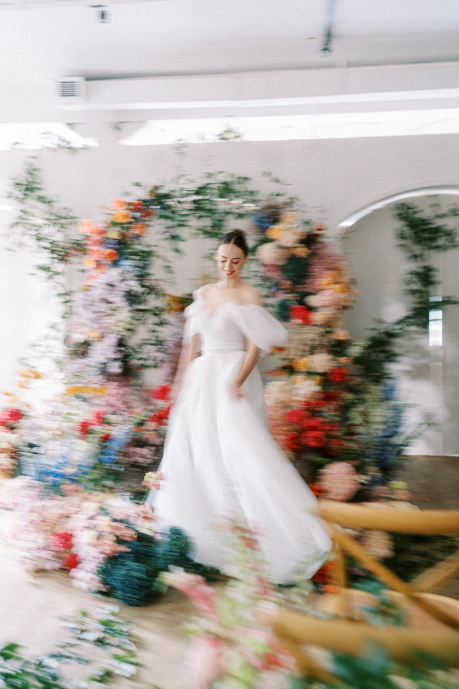 A bride in a flowing white gown surrounded by a vibrant floral archway, creating a dreamy and ethereal wedding atmosphere indoors.