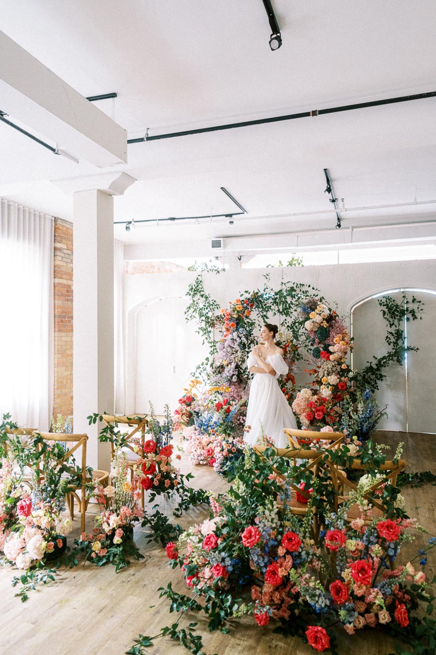 Elegant indoor wedding venue with a bride in a white gown surrounded by vibrant floral arrangements, including red, pink, and blue flowers, creating a romantic and enchanting atmosphere.