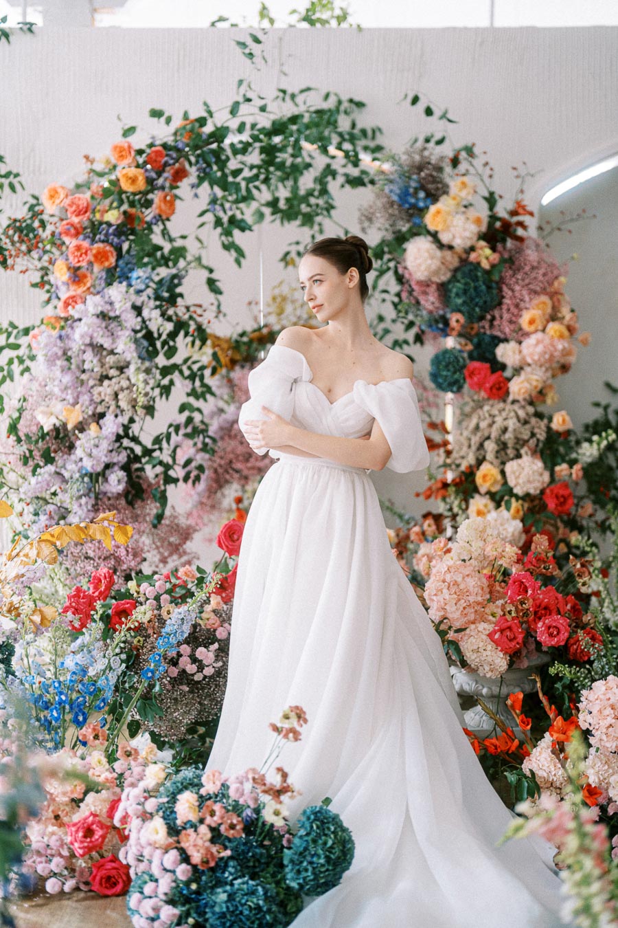 A bride in an elegant white wedding gown stands amidst a vibrant floral arrangement with various colorful flowers including pink, red, blue, and orange blooms, creating a romantic and enchanting atmosphere.