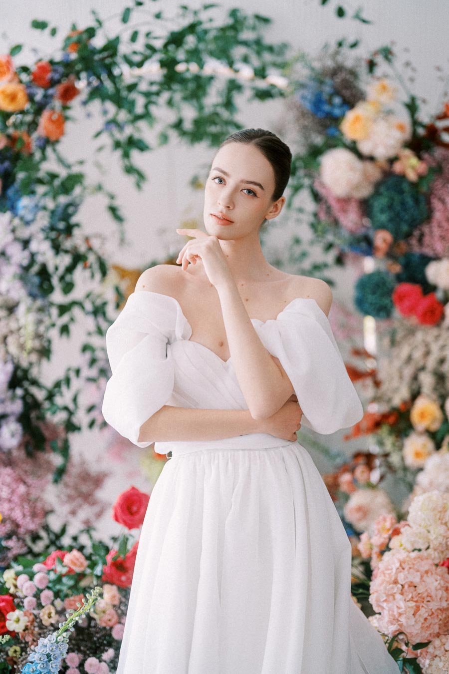 Elegant woman in a white off-shoulder dress posing amid vibrant floral background, exuding grace and style.