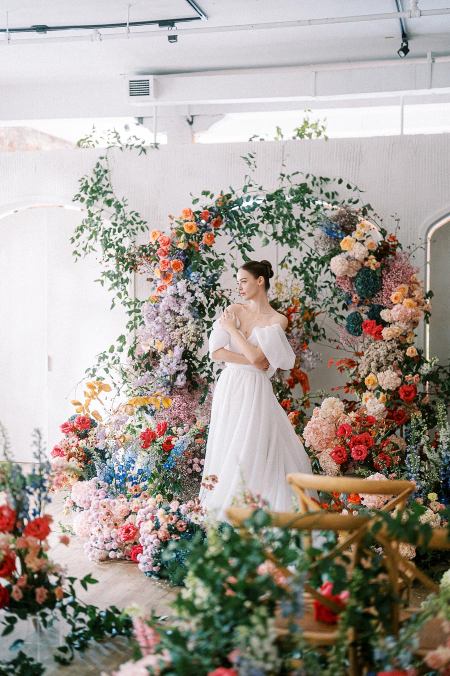 A woman in an elegant white dress stands in front of a vibrant floral arch display decorated with colorful flowers and green foliage, creating a romantic and artistic setting in a bright, modern space.