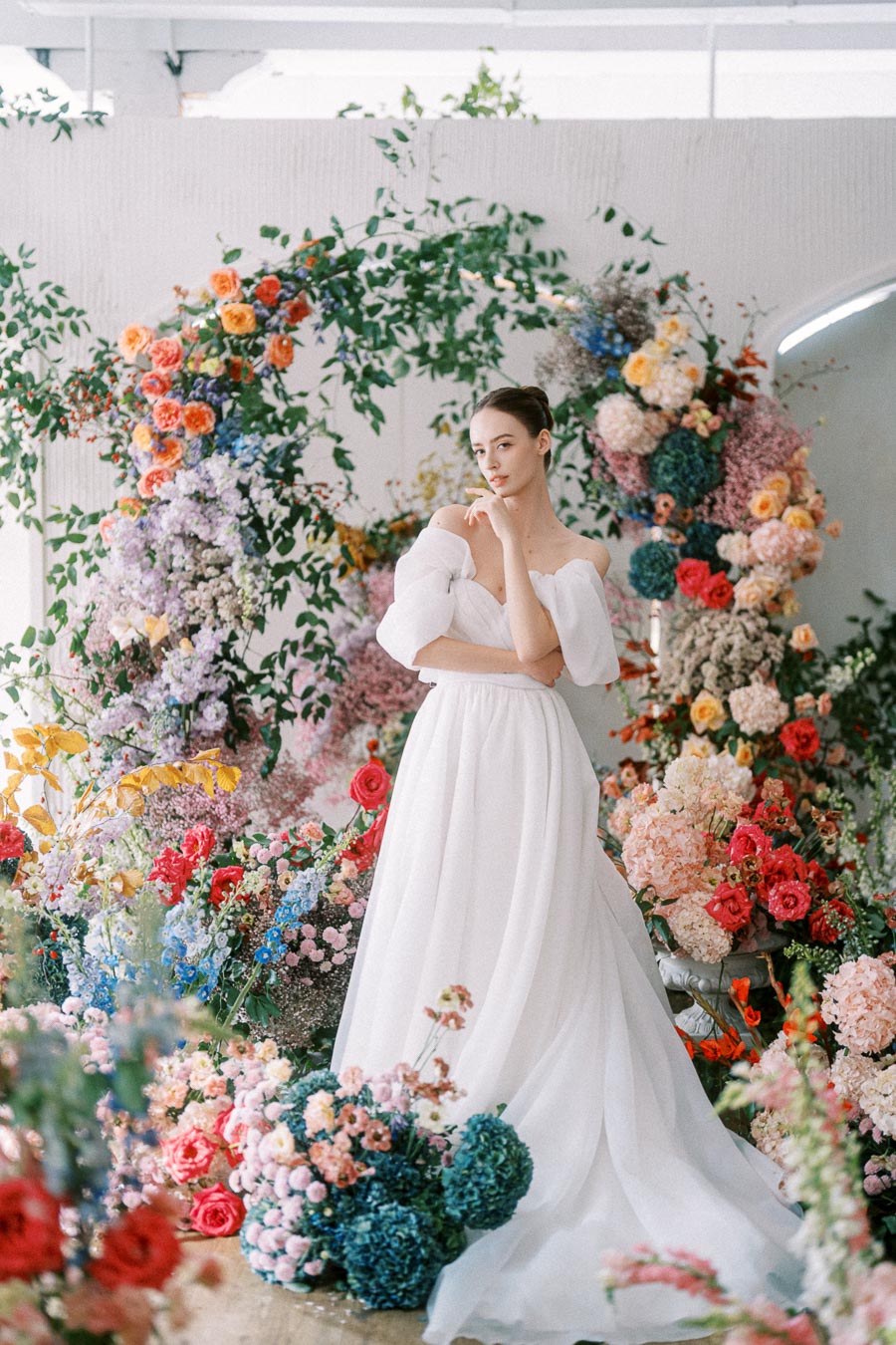 A bride in a white wedding gown stands gracefully amidst a vibrant floral arrangement with roses, hydrangeas, and greenery, creating an enchanting and romantic setting.