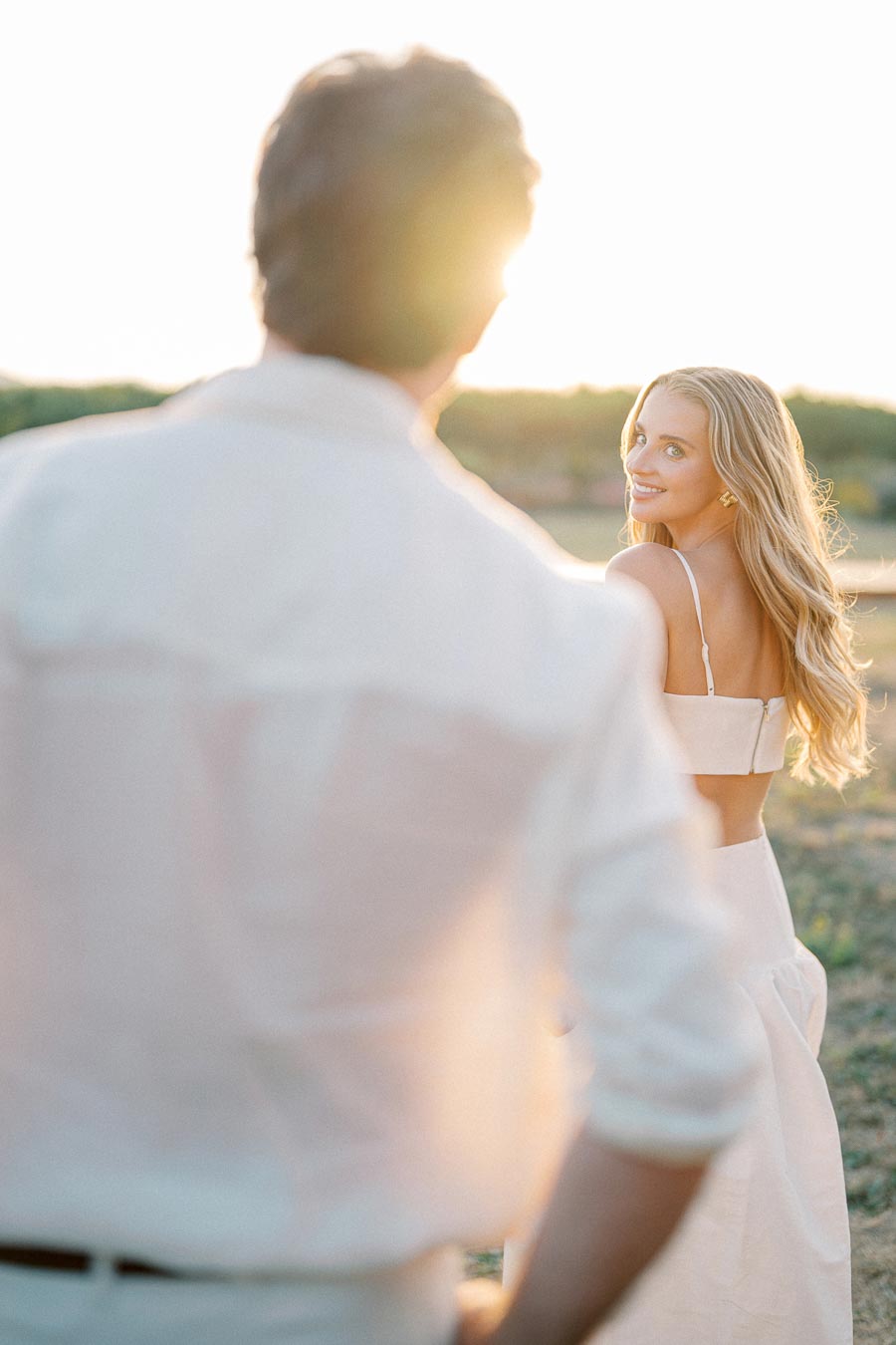 A young woman in a white dress smiles back at a man in a sunlit outdoor setting, capturing a romantic moment during golden hour.