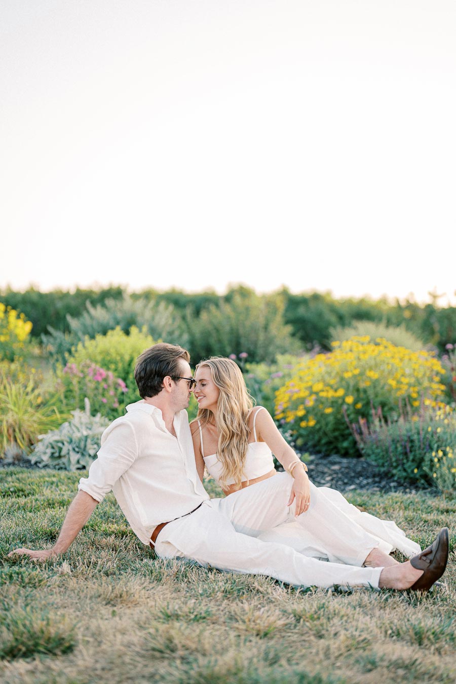 A couple in white clothing sitting on grass and leaning towards each other in a lush garden with colorful flowers, under a clear sky.