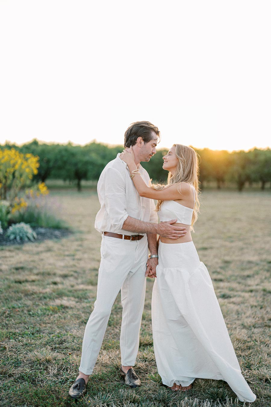 A couple in coordinated white outfits embraces lovingly in a sunlit field, surrounded by greenery and flowers, creating a romantic, outdoor atmosphere.