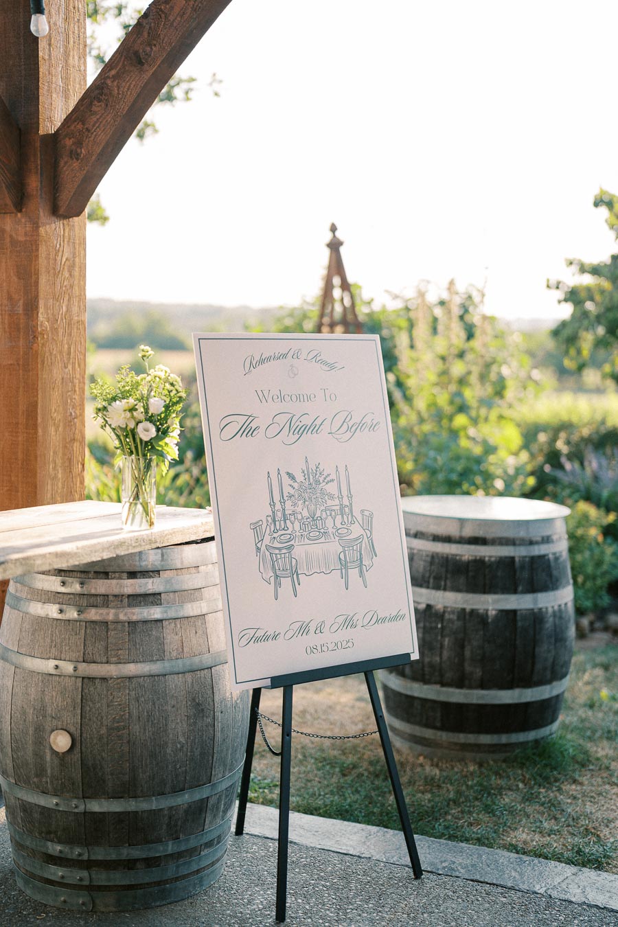 A stylish wedding welcome sign displayed outdoors on an easel, featuring elegant script text and a table setting illustration. Two wooden barrels and a vase with white flowers create a rustic yet refined atmosphere, perfect for a wedding rehearsal event.