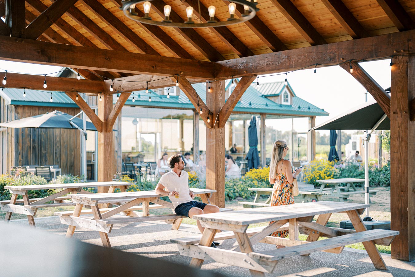 Outdoor patio with wooden picnic tables at a rustic venue, featuring a couple enjoying a sunny day under a wooden pavilion with string lights, surrounded by lush greenery and flowers.