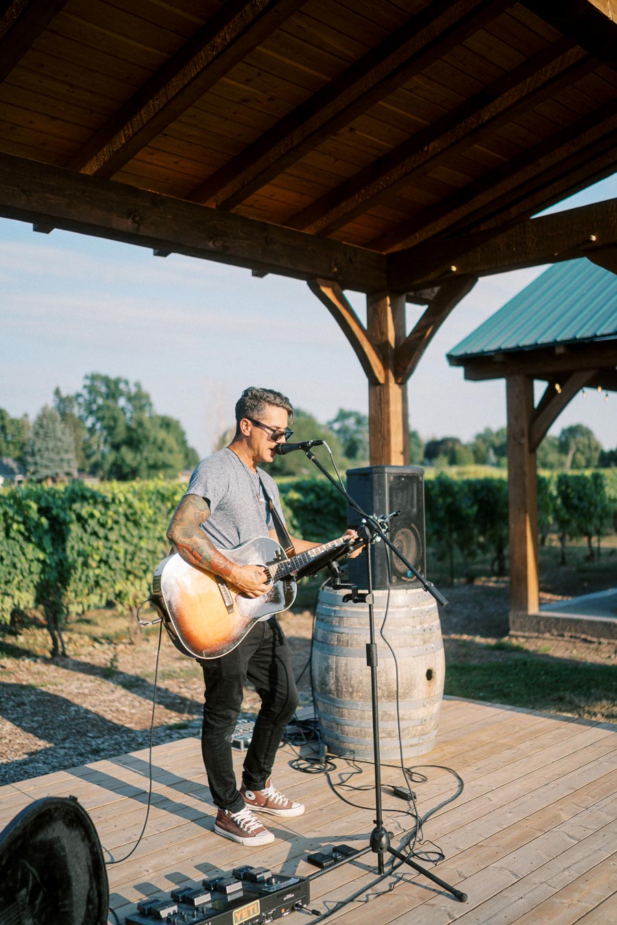 Musician performing outdoors on a wooden stage, playing an acoustic guitar, with lush green vineyard in the background, under a wooden pergola.
