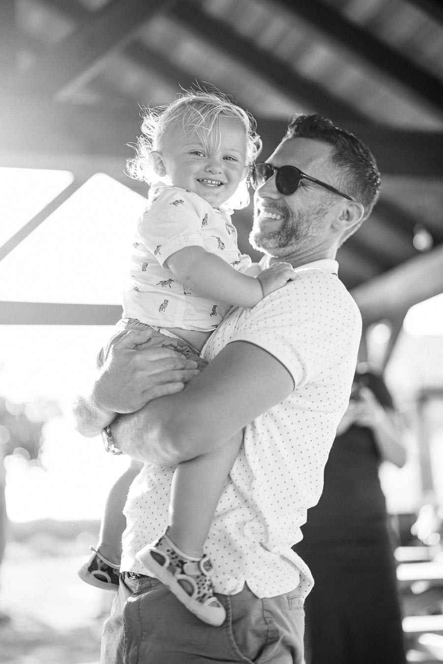 A father joyfully holding his smiling toddler in an outdoor setting, both wearing casual summer clothes, under a wooden pavilion.