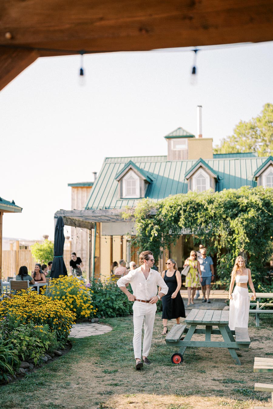 Sunny outdoor gathering at a rustic house with green roofing, surrounded by lush greenery and vibrant yellow flowers. Guests in summer attire enjoy the relaxed garden setting, featuring picnic tables and casual walking paths.