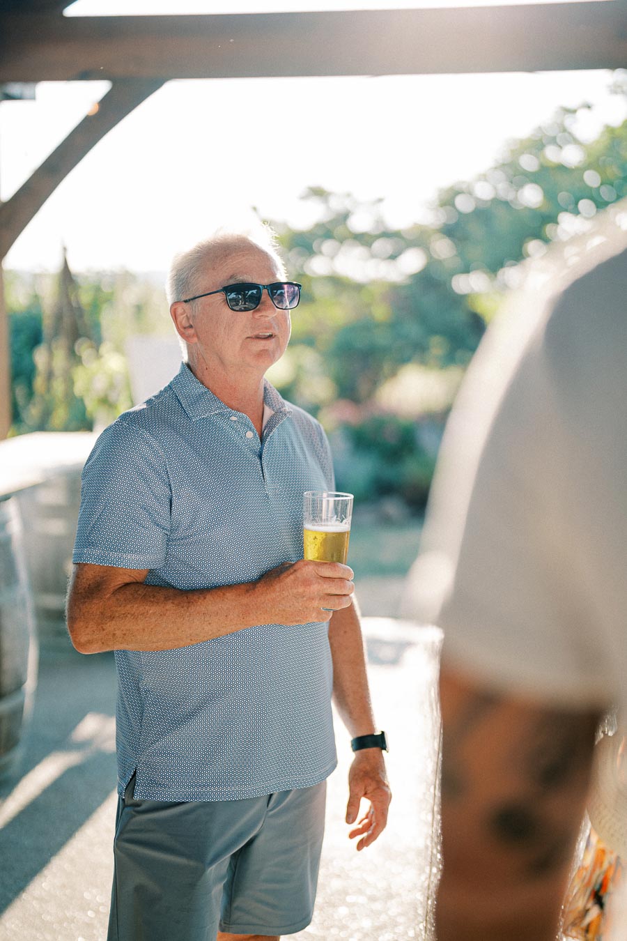 Elderly man wearing sunglasses and a blue polo shirt, holding a beer glass while enjoying a sunny outdoor gathering.