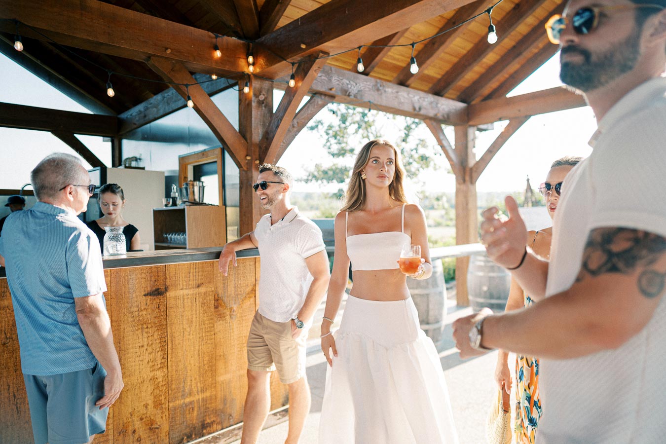 A group of people enjoying a sunny day at an outdoor bar, with a woman in a white dress holding a drink while others chat under a wooden pergola decorated with string lights.