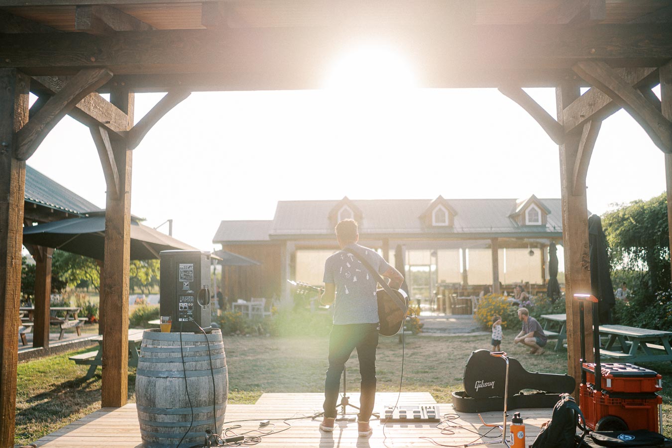 Outdoor live music performance at a rustic venue with a guitarist playing on a wooden stage at sunset, surrounded by picnic tables and lush greenery.