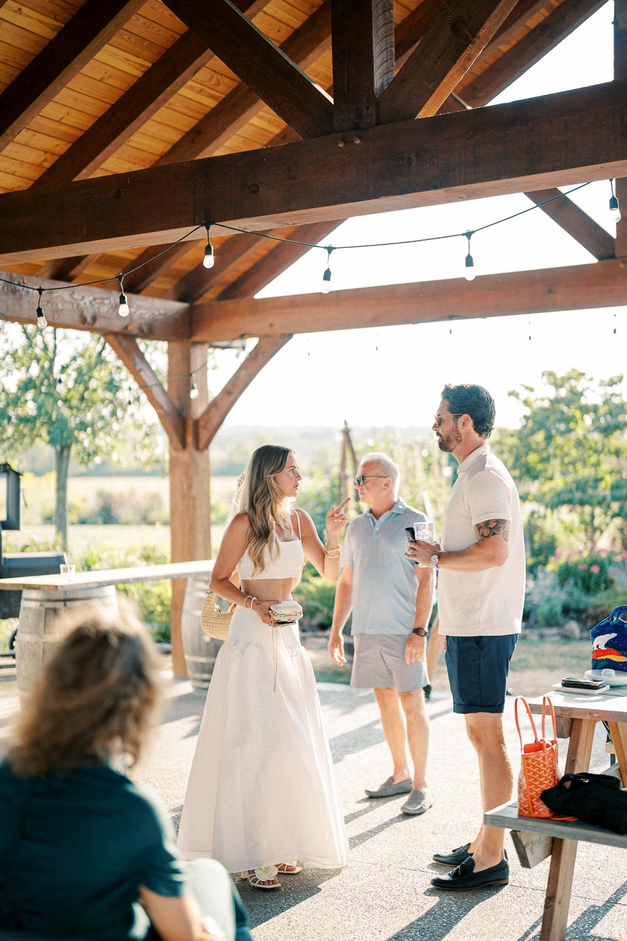 People enjoying a casual outdoor gathering under a wooden pavilion with hanging lights, surrounded by greenery and natural light.