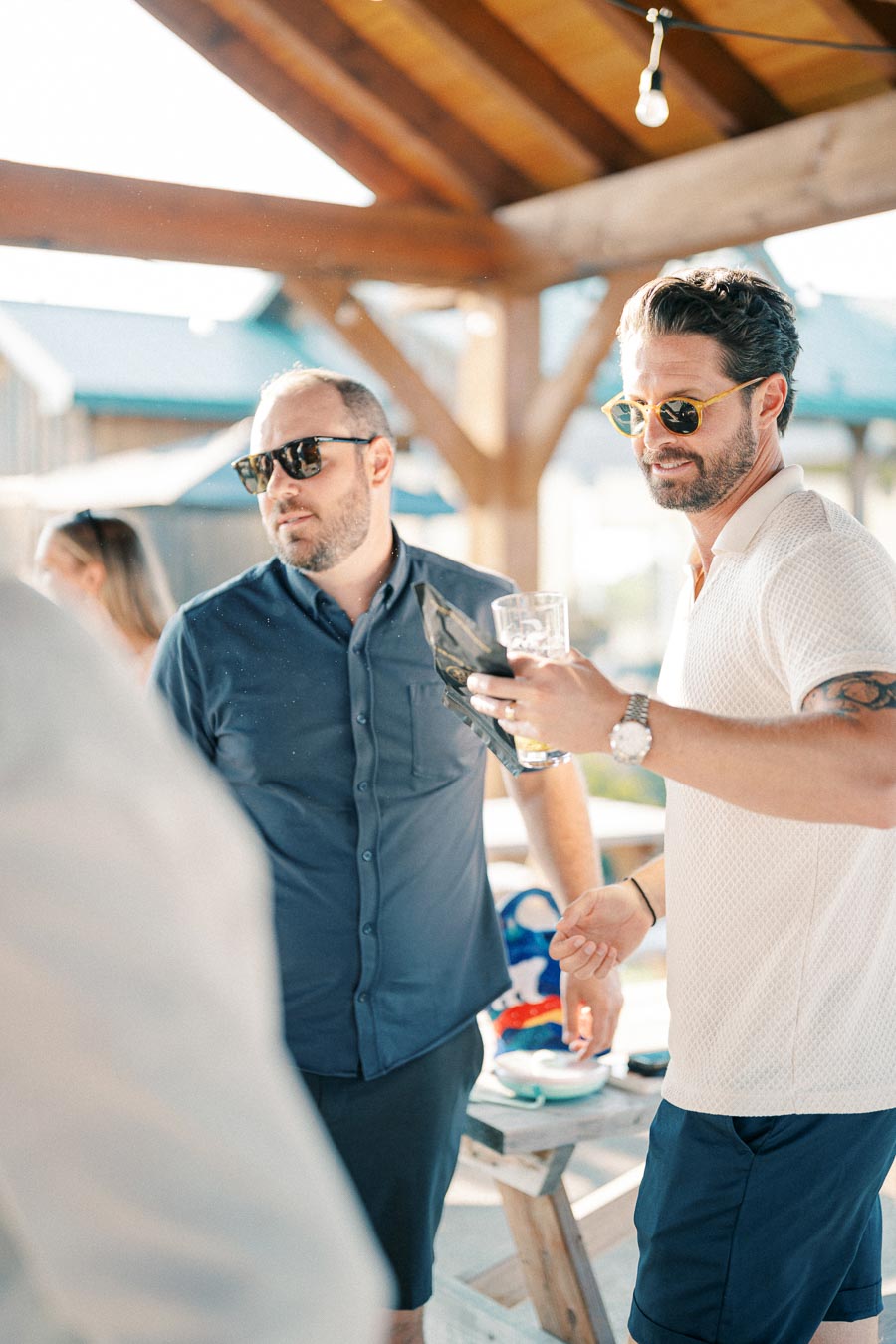 Two men wearing sunglasses enjoying a gathering under a wooden pergola, with one holding a drink and the other wearing a navy shirt in a sunny outdoor setting.