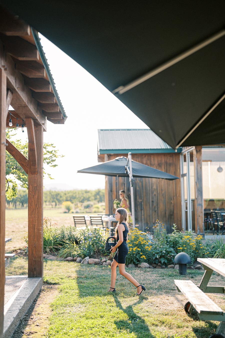 A young girl in a navy dress walks across a sunny garden patio with yellow flowers, near a rustic wooden building and outdoor seating area with large umbrellas.