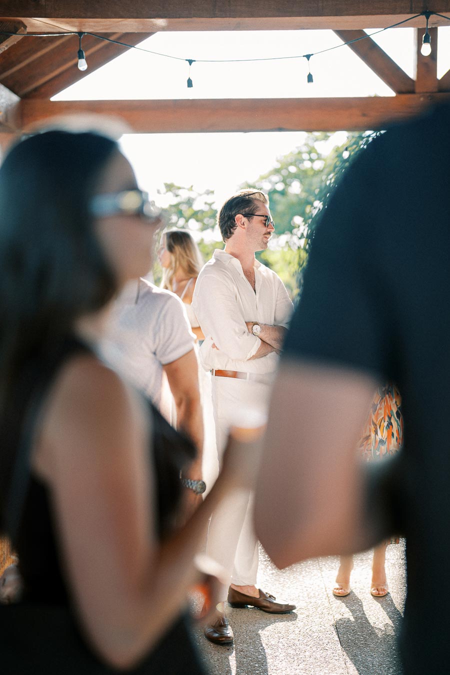 A man wearing a light-colored outfit and sunglasses stands outdoors in the sunlight, surrounded by a crowd, framed by a wooden roof and string lights.