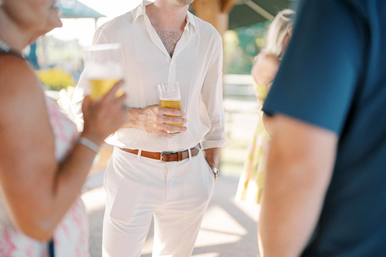People socializing at an outdoor event, holding glasses of beer, with a person in a light-colored outfit in focus in the center.