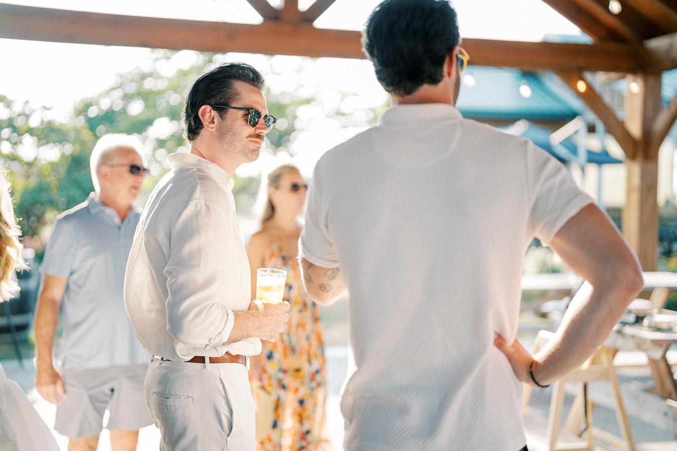 Group of people socializing outdoors under a wooden pergola, enjoying a sunny day. The individuals, casually dressed, are engaged in conversation, with one holding a drink. The background features lush greenery and soft, natural sunlight.