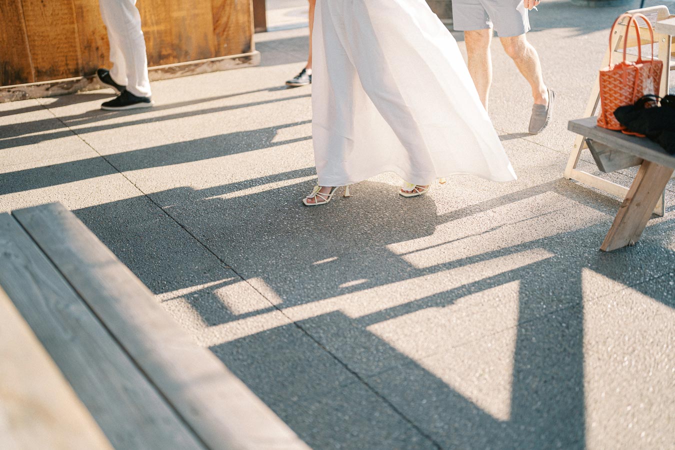 A sunlit outdoor setting featuring the lower half of people walking on pavement, showcasing a person in a white flowing dress and heels, accompanied by another in casual shorts. Nearby, a wooden bench holds an orange handbag with sunny shadows falling across the scene.
