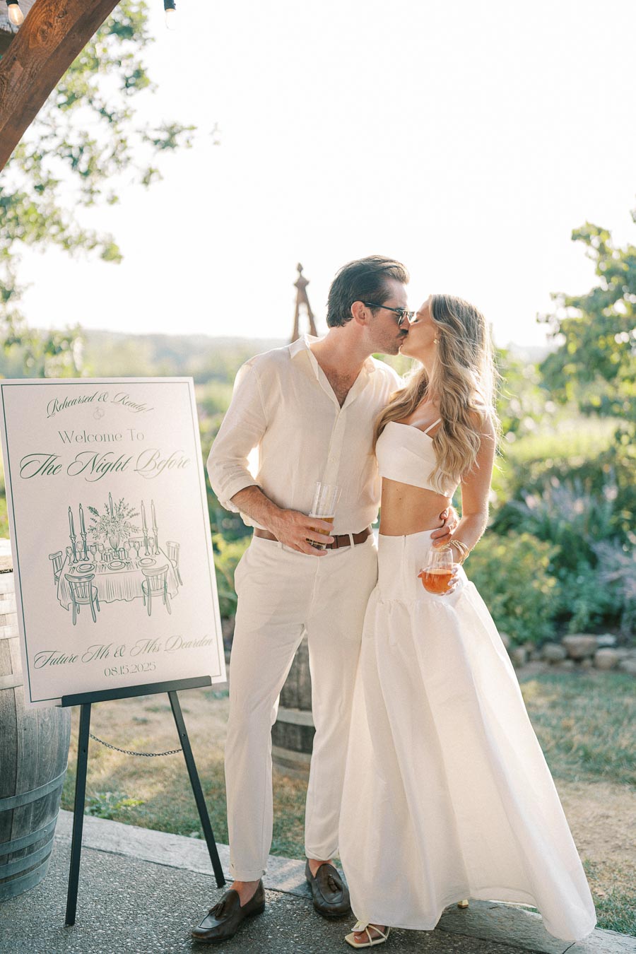 A couple wearing white outfits shares a romantic kiss next to a wedding sign, set in an outdoor garden venue under a soft, natural sunlight.