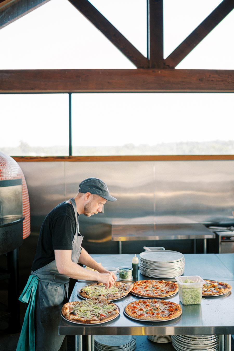 A chef in a modern kitchen prepares multiple freshly baked pizzas on a steel countertop, with stacks of plates and ingredients visible, creating a vibrant culinary atmosphere.