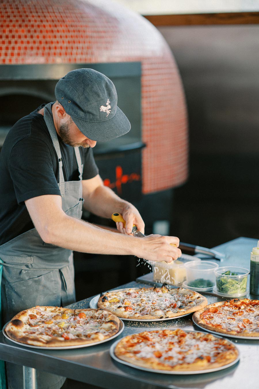 Chef grating cheese over freshly baked wood-fired pizzas in a rustic kitchen setting.