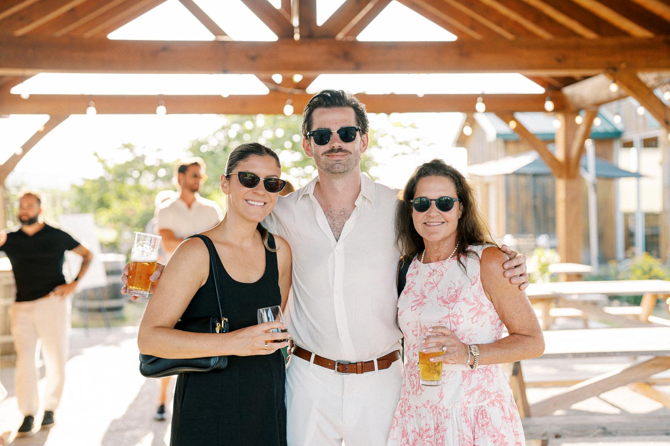 Group of three friends wearing sunglasses, enjoying beverages at an outdoor gathering under a wooden pavilion, with a warm, sunny backdrop.