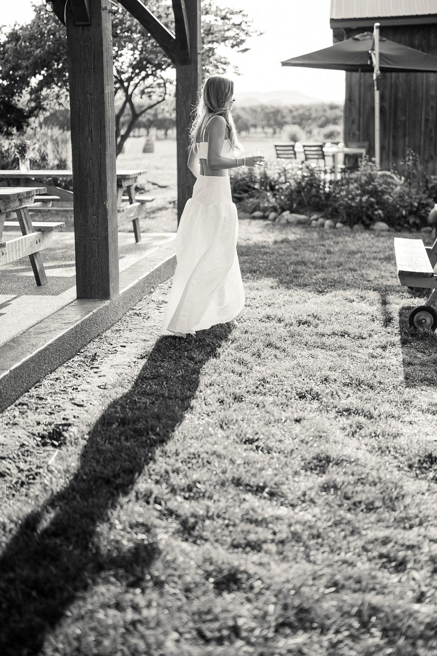 Black and white photo of a woman in a flowing dress walking outside near a wooden picnic area and barn, casting a long shadow on the grass.
