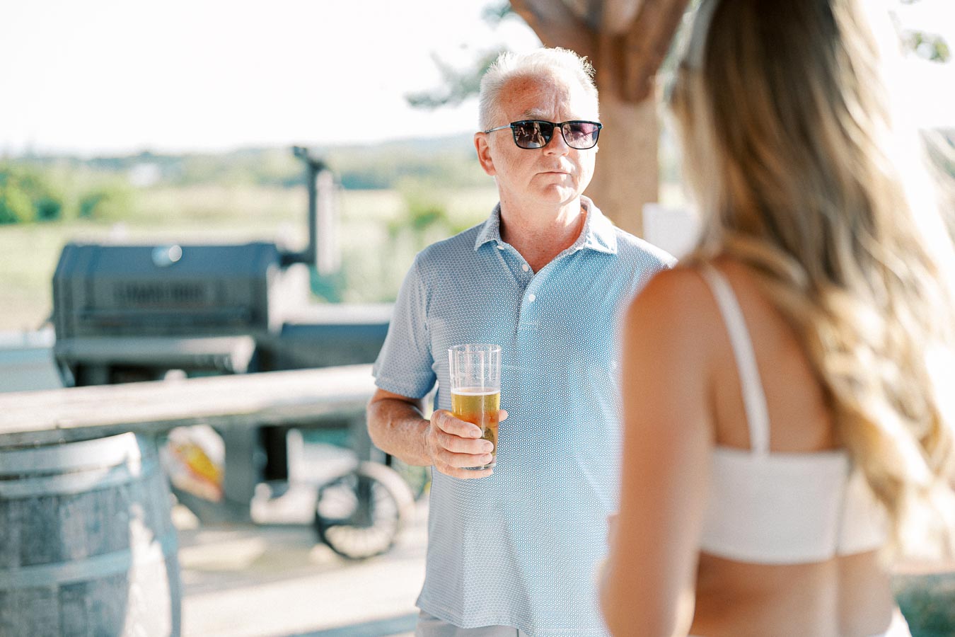 Man enjoying a drink outdoors in sunny weather, wearing sunglasses and a blue polo shirt, while conversing with a woman in a white top.