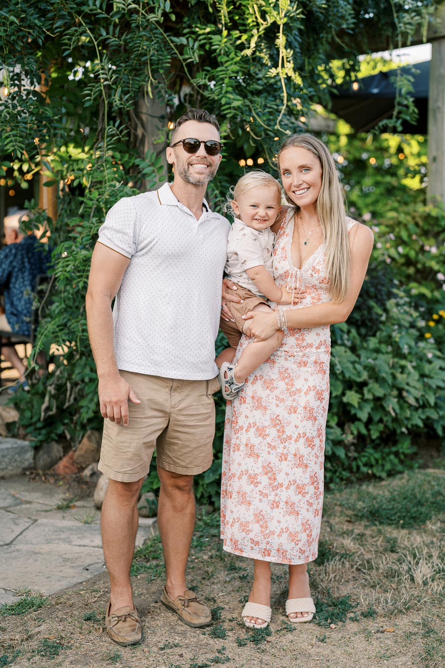 Happy family posing outdoors in a garden with lush greenery. A man wearing sunglasses and a white polo shirt stands beside a woman in a floral dress holding a smiling toddler.
