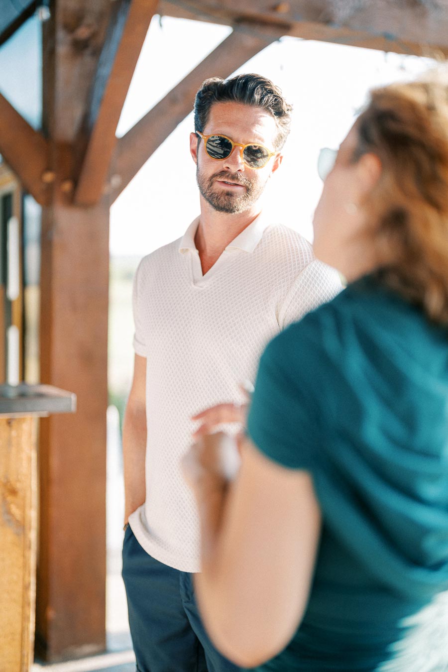 A man wearing sunglasses and a white polo shirt stands outdoors, engaging in a conversation with a woman in a teal shirt, with wooden beams and sunlight in the background.