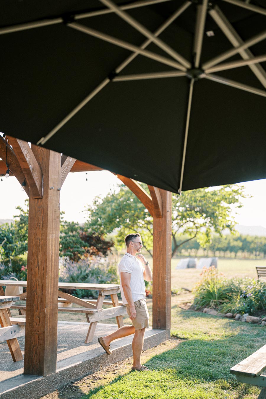 A man in a casual summer outfit stands under a large patio umbrella, enjoying a sunlit garden with wooden picnic tables and vibrant greenery.