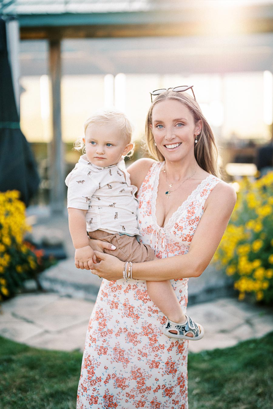 Woman in a floral dress holding a toddler, standing in a garden with yellow flowers and sunlight in the background.