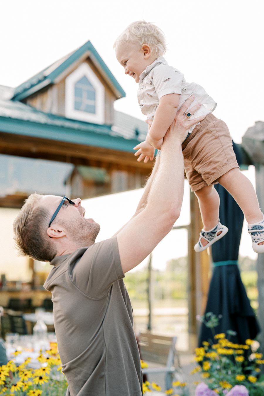 A father joyfully lifting his smiling toddler son outdoors near a house with a garden in the background, surrounded by yellow flowers.