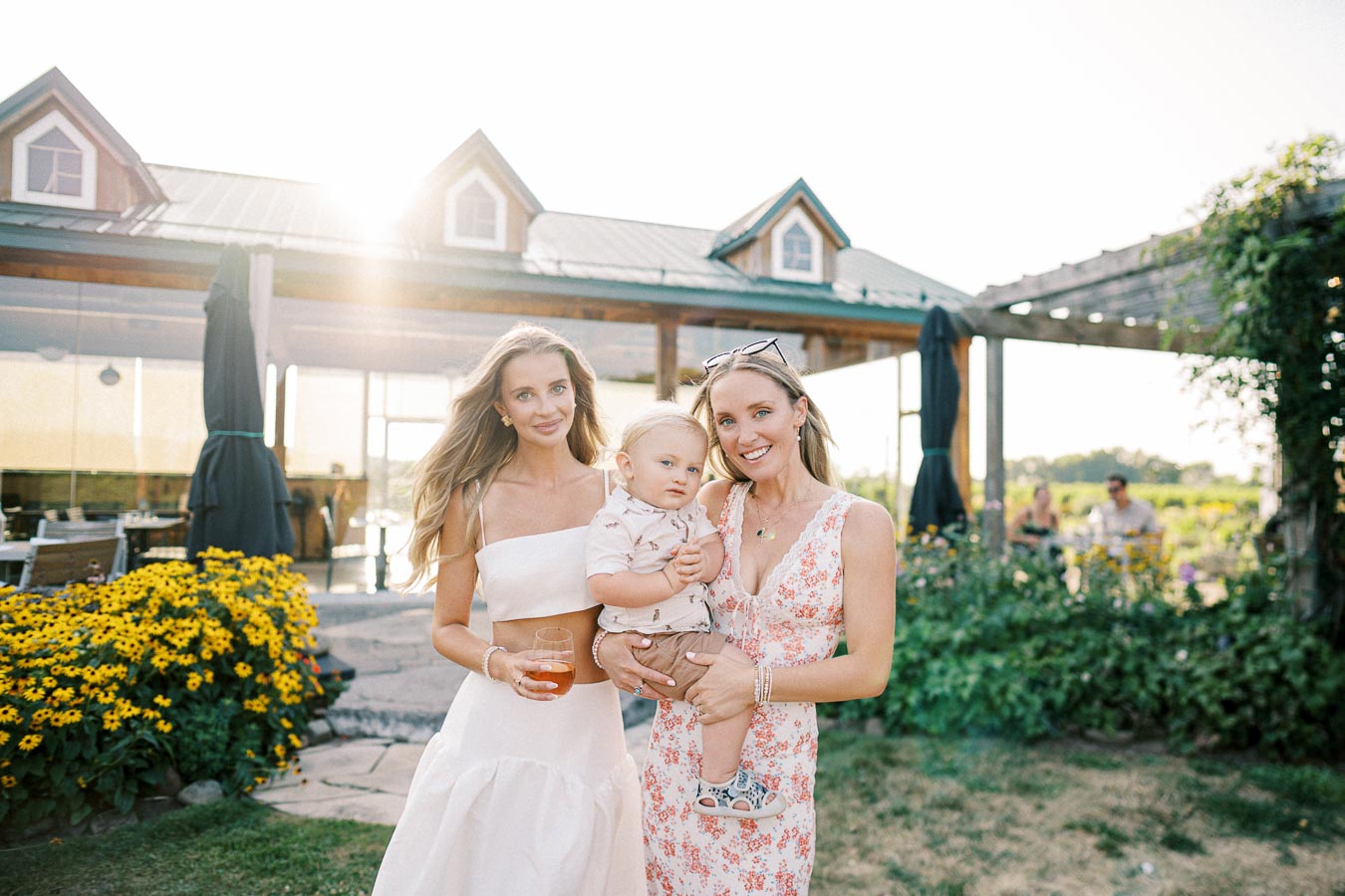Two women and a child stand in a sunlit garden with a rustic building in the background. The women wear summer dresses, and one holds a glass of juice, while the other holds the child. Yellow flowers and greenery surround them, creating a warm, inviting atmosphere.