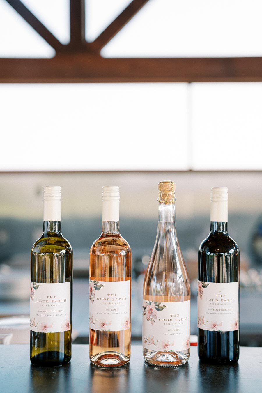 Four bottles of wine from The Good Earth winery displayed on a countertop, featuring a variety of wines including white, rosé, sparkling, and red, with floral labels.