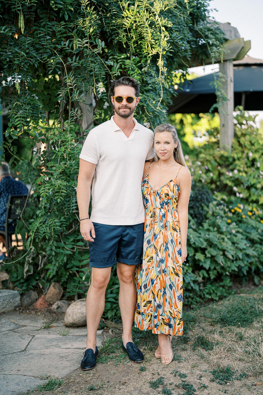 A couple posing outdoors in summer attire, standing in front of lush greenery and a garden setting. The man is wearing a light-colored shirt with shorts, and sunglasses, while the woman is in a vibrant floral dress.