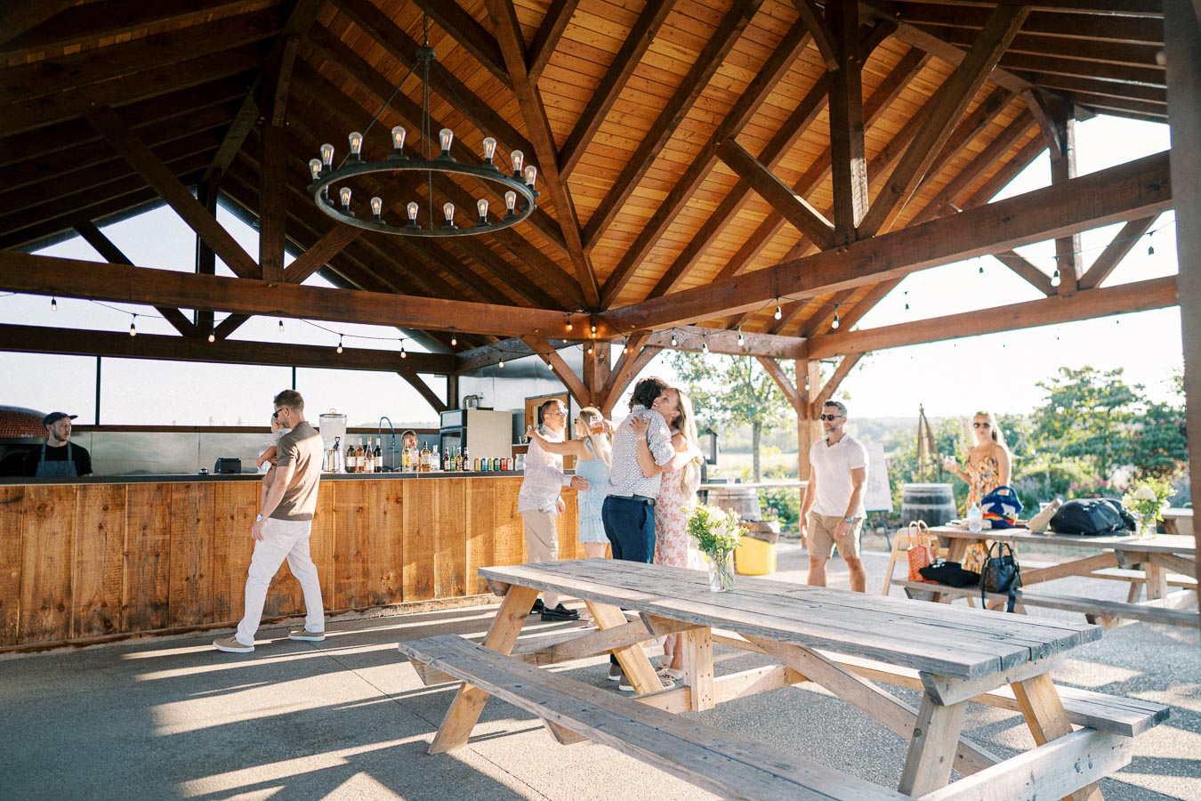 Outdoor social gathering at a wooden pavilion with people enjoying drinks and conversation. Rustic decor, picnic tables, and a bartender serving beverages under a chandelier, creating a warm and inviting atmosphere.