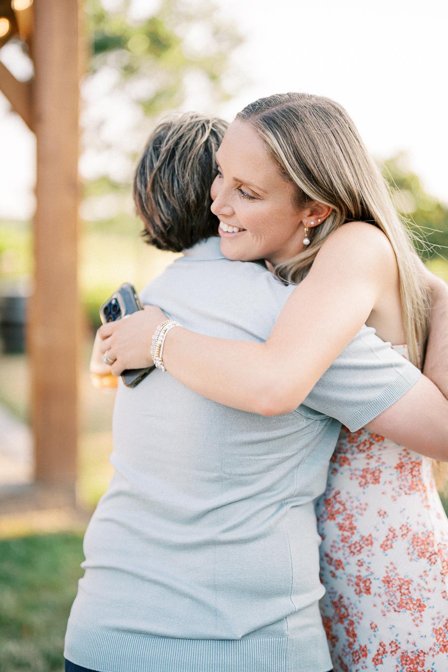 A woman with long hair smiling while hugging another person outdoors, wearing a floral dress and holding a smartphone.